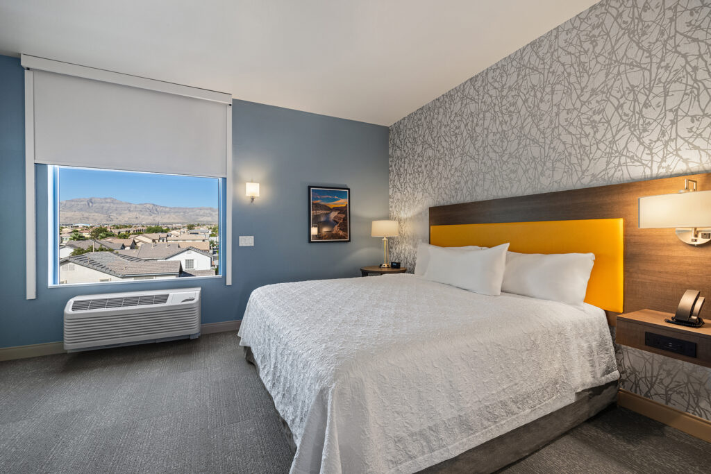 A modern hotel room at Home2 Suites by Hilton features a large bed with white bedding and a yellow headboard. The walls boast a blue, patterned design, while the window offers a stunning mountain view. An air conditioner sits below the window, and there's a lamp on the side table.