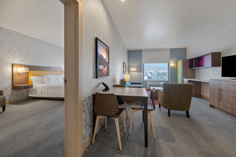 A modern Home2 Suites by Hilton hotel room offers a cozy sitting area, a table with chairs, and a TV. The bedroom boasts a large bed and nightstand. A window frames outdoor views, enhancing the room's spacious and well-lit interior.