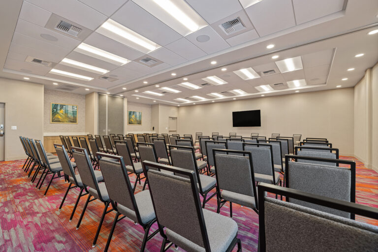 A conference room with rows of gray chairs facing a large screen, reminiscent of the sleek design found at Home2 Suites by Hilton. The vibrant red and orange carpet adds a touch of warmth, while two framed artworks and recessed lighting complete the modern ambiance.