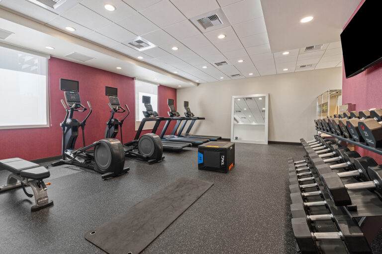 A modern gym room with exercise equipment, including treadmills, ellipticals, a weight bench, dumbbells, and a large mirror on the wall. The room has maroon walls and a black rubber floor. Ceiling lights are evenly spaced, illuminating the area.