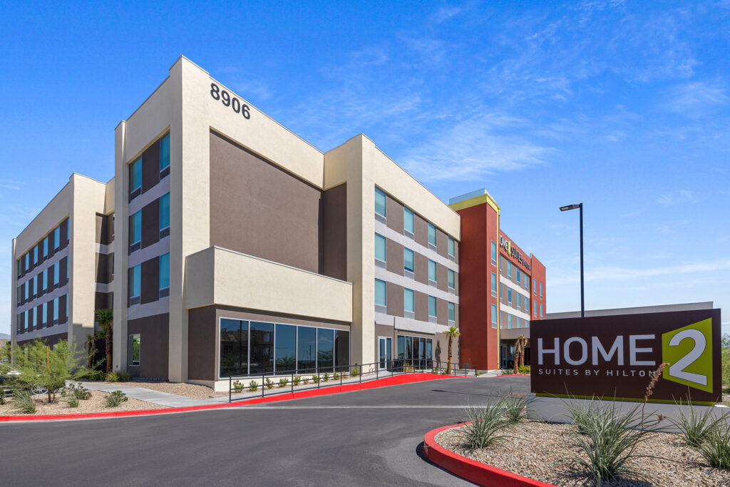 A modern home2 suites hotel building with multiple floors and large windows, featuring brown, beige, and red accents. The hotel sign reads Home2 Suites by Hilton. The surrounding area has landscaped rocks and minimal plant life under a clear blue sky.