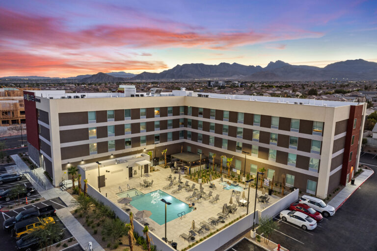 A modern Home2 hotel with a central courtyard featuring a swimming pool and patio seating. The surrounding landscape includes mountains under a vibrant sunset sky, with parked cars in the foreground.
