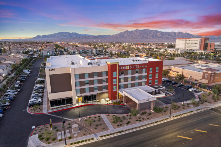 Aerial view at dusk reveals the modern architecture of Home2 Suites by Hilton in Nevada. The four-story hotel, adorned in red, brown, and white hues, is encircled by a spacious parking lot with scenic mountains providing a stunning backdrop.