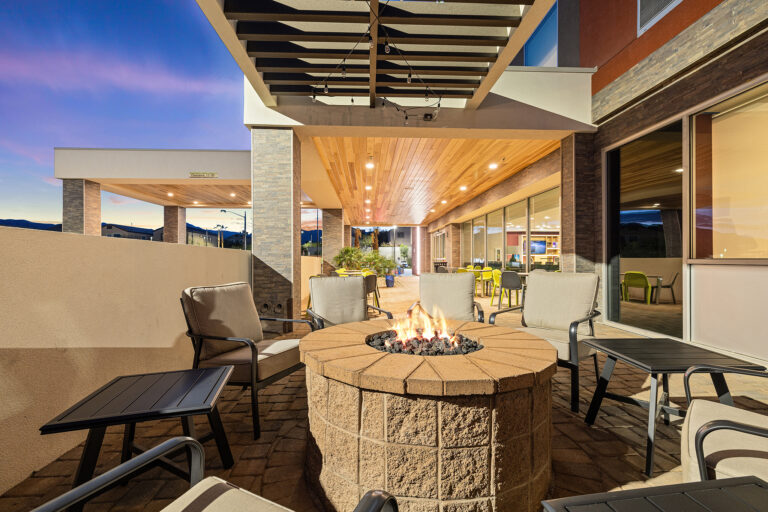 The outdoor patio at Home2 Suites Las Vegas features cushioned chairs surrounding a stone fire pit. The area is covered by a wooden ceiling and illuminated by warm lights, with the clear Nevada evening sky providing a picturesque backdrop.