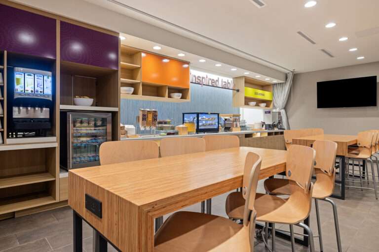 The modern dining area at Home2 Suites in Las Vegas features wooden tables and chairs, complemented by beverage dispensers, snack displays, and a fridge. A flat-screen TV adorns the wall, while bright lighting and colorful accents enhance the welcoming Hilton atmosphere.