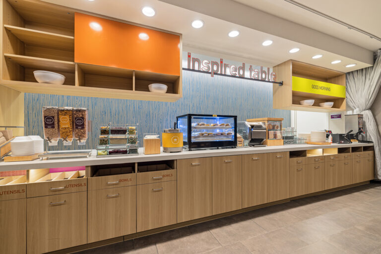 A modern breakfast buffet area at Home2 Suites by Hilton in Las Vegas features wooden cabinets and shelves, cereal dispensers, a refrigerated display with pastries, and a countertop with stacked bowls and utensils. Bright lighting and an Inspired Table sign enhance the inviting atmosphere.