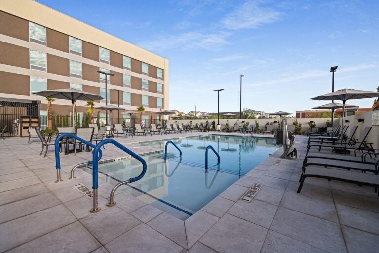 The outdoor hotel swimming pool at Home2 Suites in Las Vegas features crystal-clear water, surrounded by lounge chairs and umbrellas. A modern building with shimmering windows stands in the background against a clear blue Nevada sky, enhancing the relaxing atmosphere.