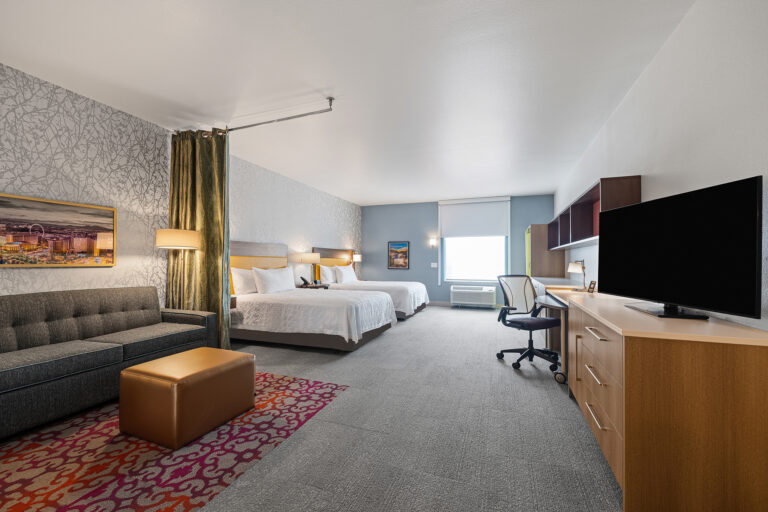 Experience the spacious comfort of our Hilton home2 suites hotel room featuring two queen beds with wooden headboards, a gray sofa, and a brown ottoman. A desk with a chair and flat-screen TV complete this stylish space, while artwork adorns the walls and large windows let in abundant natural light.