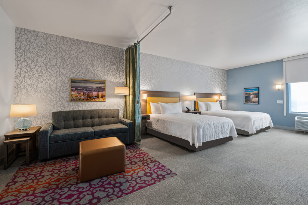 A modern hotel room at Hilton, featuring two queen beds, a dark blue sofa, a brown ottoman, and colorful wall art. The room is enhanced by soft lighting, a gray and white patterned accent wall, and a small side table with a lamp.