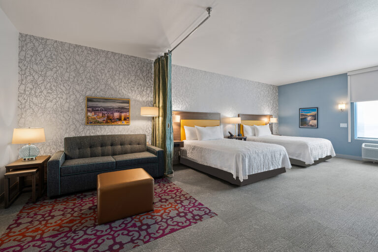 A modern hotel room at Hilton, featuring two queen beds, a dark blue sofa, a brown ottoman, and colorful wall art. The room is enhanced by soft lighting, a gray and white patterned accent wall, and a small side table with a lamp.