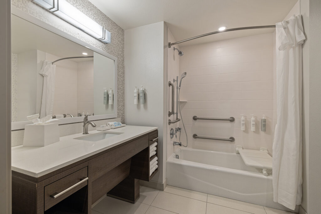 A modern ada bathroom at Home2 Suites features a large mirror above a sink on a wooden vanity. The space includes a bathtub with a shower curtain, mounted shampoo bottles, and a foldable seat. White towels are neatly stacked on the vanity shelf for your convenience.