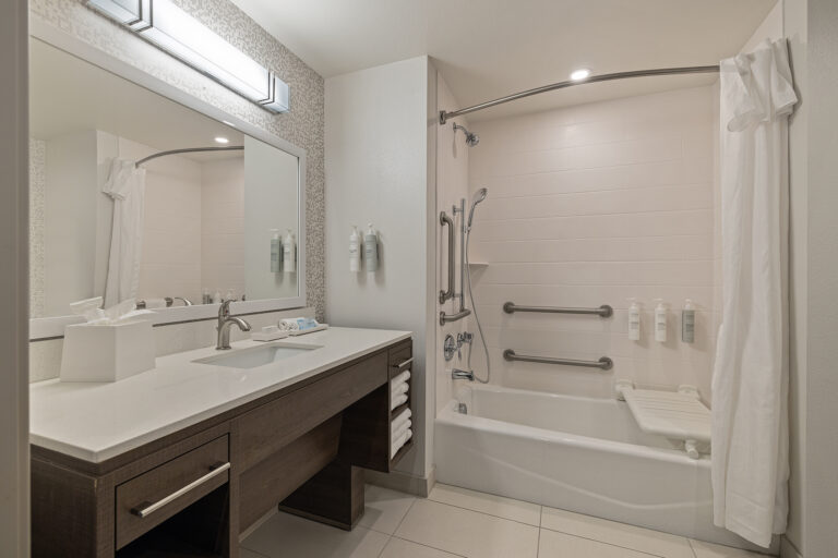 A modern ada bathroom at Home2 Suites features a large mirror above a sink on a wooden vanity. The space includes a bathtub with a shower curtain, mounted shampoo bottles, and a foldable seat. White towels are neatly stacked on the vanity shelf for your convenience.