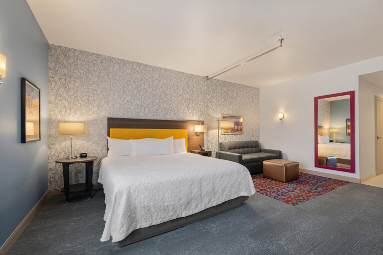 A modern hotel room at Home2 Suites by Hilton features a large bed with a yellow headboard, a gray sofa, and a patterned area rug next to a brown ottoman. The decorative wallpaper and red-framed mirror, combined with ambient lighting, create a cozy atmosphere.
