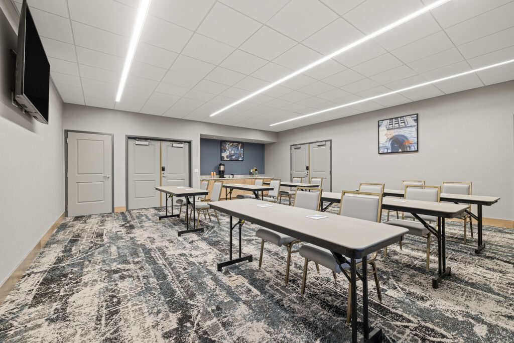 A modern conference room at the Homewood Suites by Hilton in Wisconsin boasts light gray walls and a ceiling with fluorescent lights. It features long tables surrounded by chairs, a wall-mounted TV, and framed artwork above a patterned carpet.