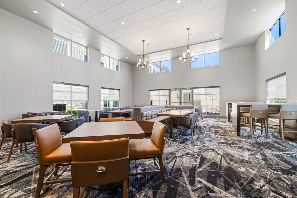 The spacious modern lounge at Homewood Suites in Milwaukee, Wisconsin, boasts high ceilings and ample natural light. It features brown leather chairs, wooden tables, and a patterned carpet. Chandeliers hang from above, with large windows lining the walls to let sunlight pour into the room.