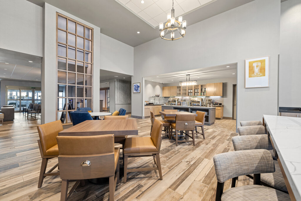 Spacious modern dining area with wooden tables and cushioned chairs, featuring a central chandelier. The open space leads to a buffet bar with hanging lights. Light wood flooring complements neutral wall colors, enhanced by decorative art on the walls—an elegant retreat in Hilton Milwaukee.