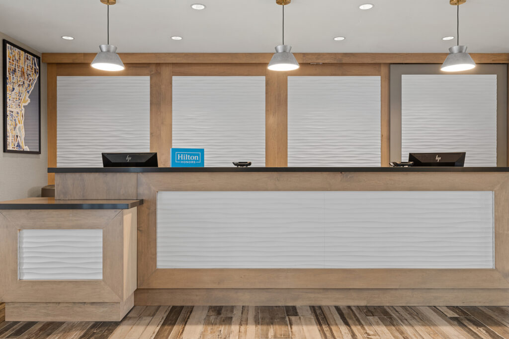 A modern hotel reception area with a wooden desk and white paneling. Two computer monitors sit beneath an elegant blue Hilton sign, reflecting the charm of Milwaukee. Pendant lights hang from the ceiling, illuminating a framed map of Wisconsin on the wall to the left.