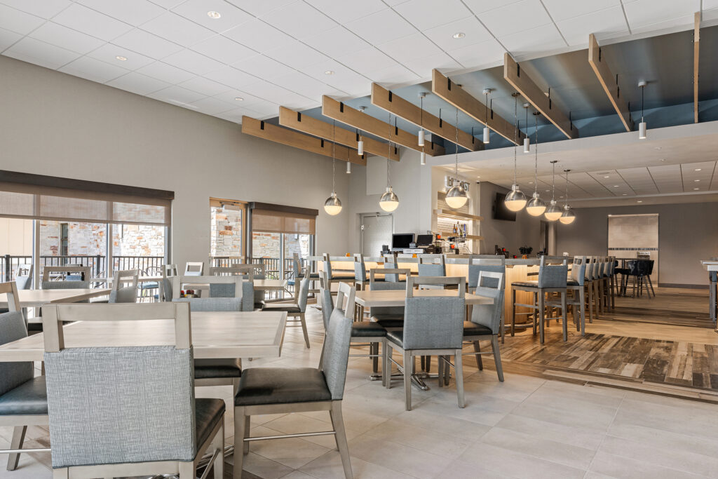 A modern, spacious dining area at the Hilton features grey chairs and light wooden tables. The ceiling showcases wooden beams and hanging pendant lights. Large windows allow natural light, with a bar area in the background, providing a welcoming atmosphere for guests to enjoy in Milwaukee.
