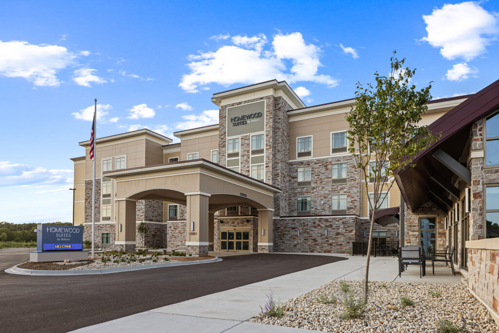 The image showcases a Homewood Suites by Hilton in Wisconsin with a charming stone facade, blue sky, and scattered clouds. An inviting covered entrance, fluttering American flag, trees, and a "Welcome" sign add warmth to the scene, all encompassed by a paved driveway and walkway.