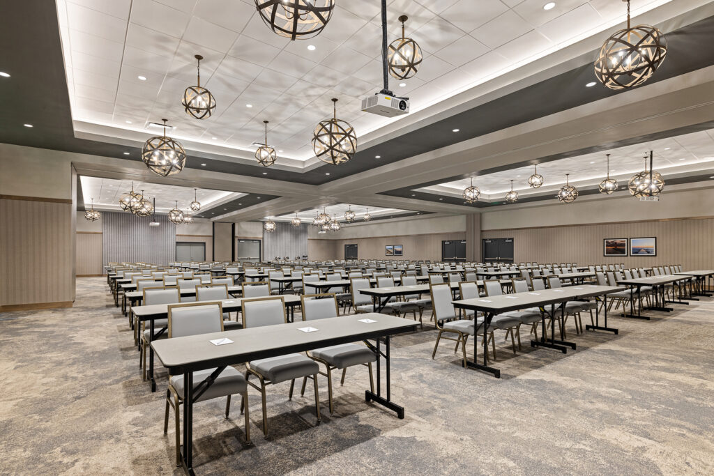 A spacious, modern conference room at the Milwaukee Hilton, featuring rows of tables and chairs facing a drop-down screen. The room is well-lit with spherical chandeliers, and the carpet displays a neutral pattern. Perfect for your next event or meeting at Homewood Suites.