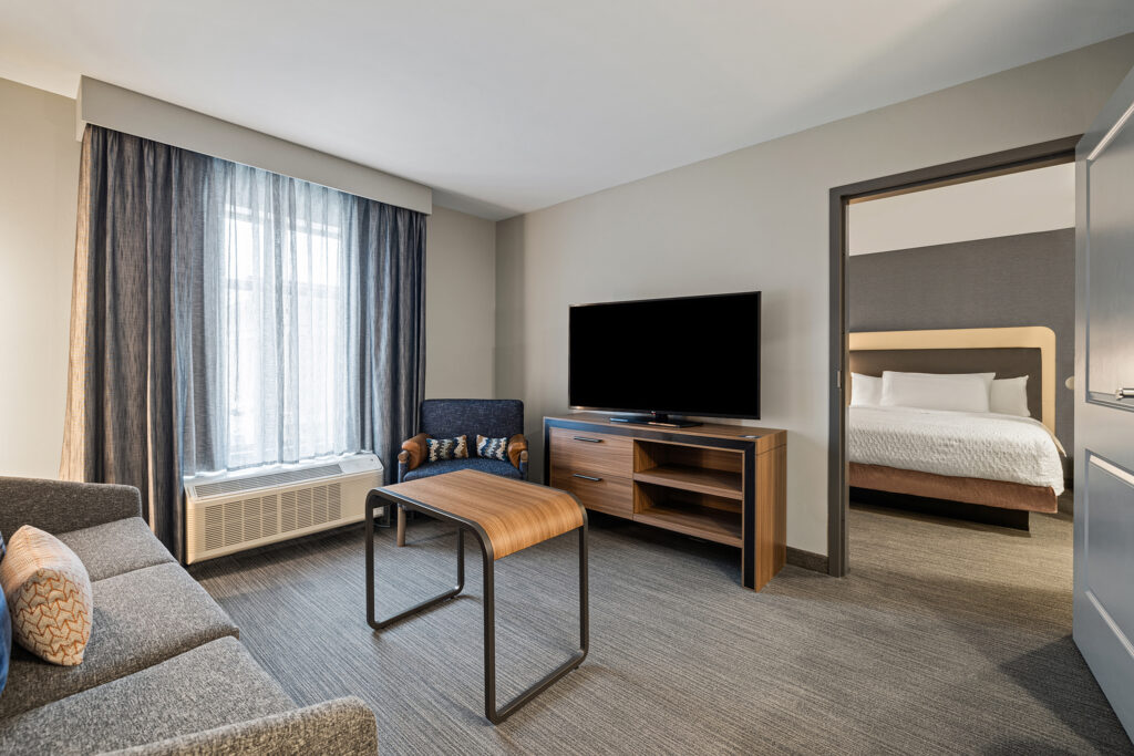Experience the modern comforts of our Hilton suite, featuring a stylish living area with a sofa, chair, and coffee table. A flat-screen TV stands ready for entertainment. The bedroom boasts a neatly made bed, while large windows invite natural light into this Wisconsin retreat.