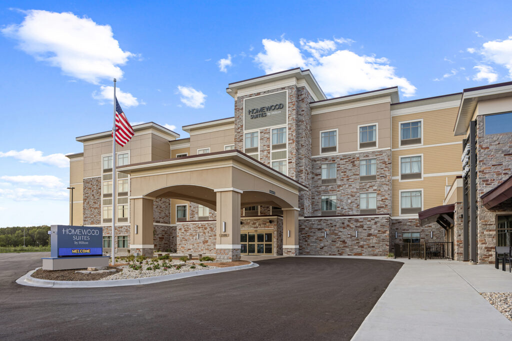 A four-story hotel building with a beige and stone exterior stands proudly in Wisconsin. The entrance features a covered driveway adorned with an American flag, while a blue sign displaying Homewood Suites by Hilton is prominent. The sky is clear with a few clouds, completing the picturesque scene.