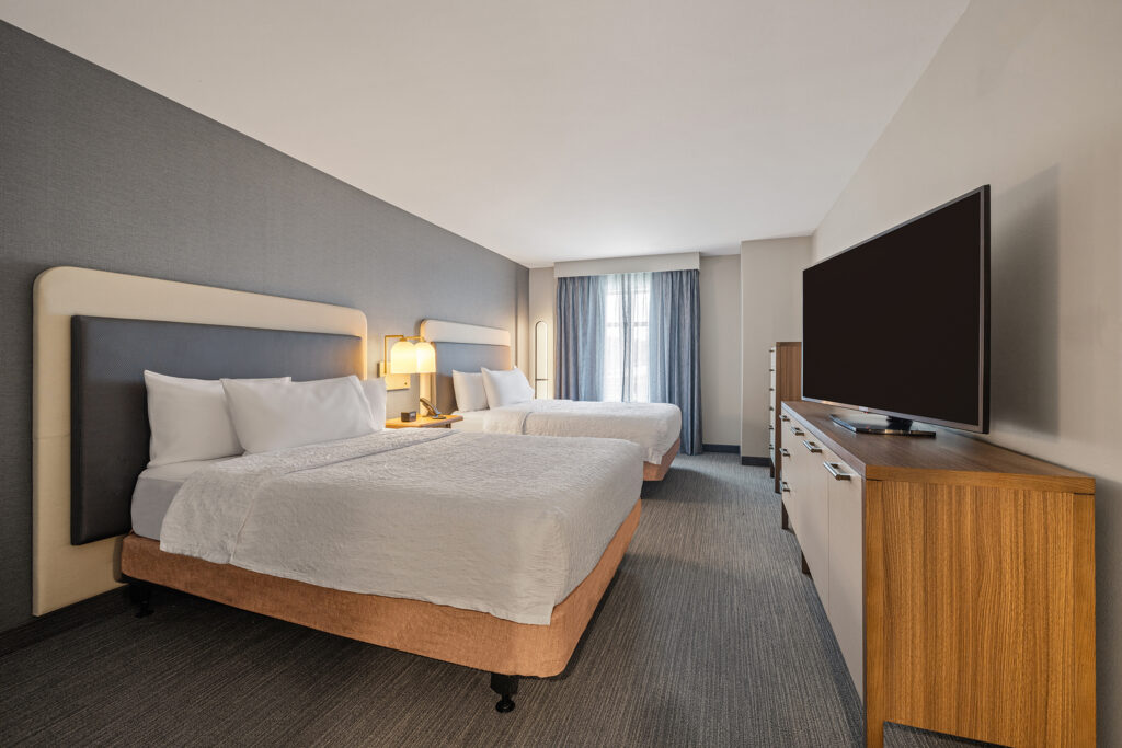 This inviting Homewood Suites by Hilton hotel room features two neatly made double beds, a wooden dresser, and a large flat-screen TV. A window with sheer curtains lets in ample natural light, while a small nightstand with a lamp is perfectly placed between the beds for added convenience.