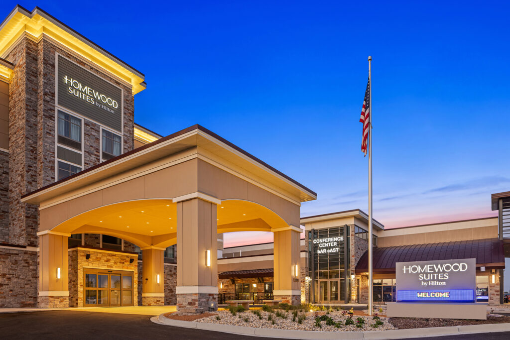Exterior of a Homewood Suites by Hilton hotel during twilight in Milwaukee, Wisconsin. The building features stone and beige accents with a covered entrance. An American flag is flying near the entrance, and a lit sign displays the hotel name and welcome message.