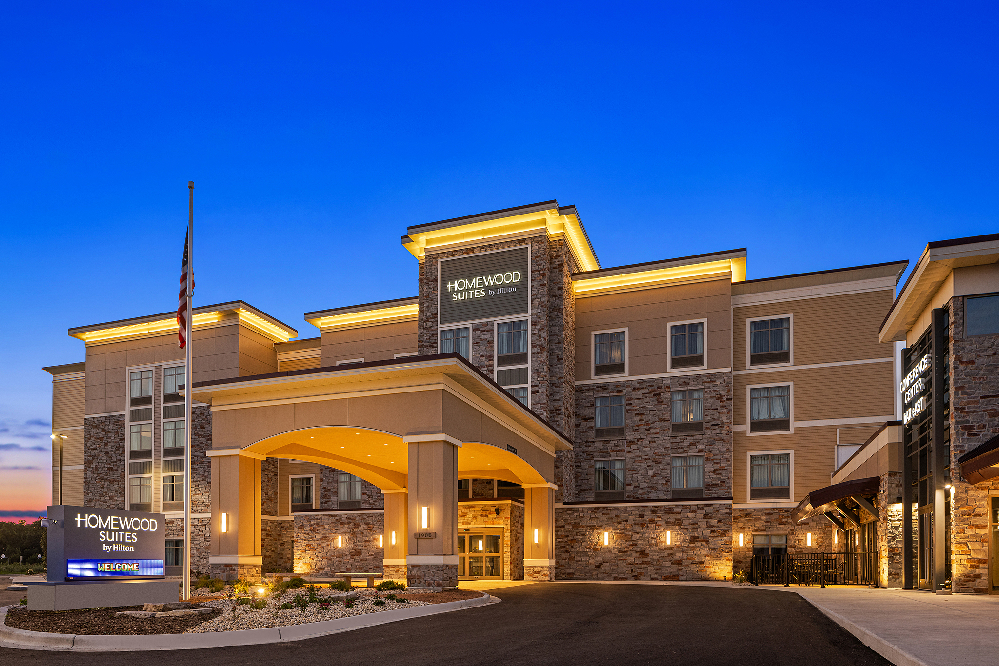 Exterior view of a Homewood Suites hotel by Hilton in Milwaukee at dusk. The building features a well-lit entrance with a covered driveway. The façade combines beige siding and stone accents. An American flag is visible on the left, set against Wisconsin's deep blue sky.
