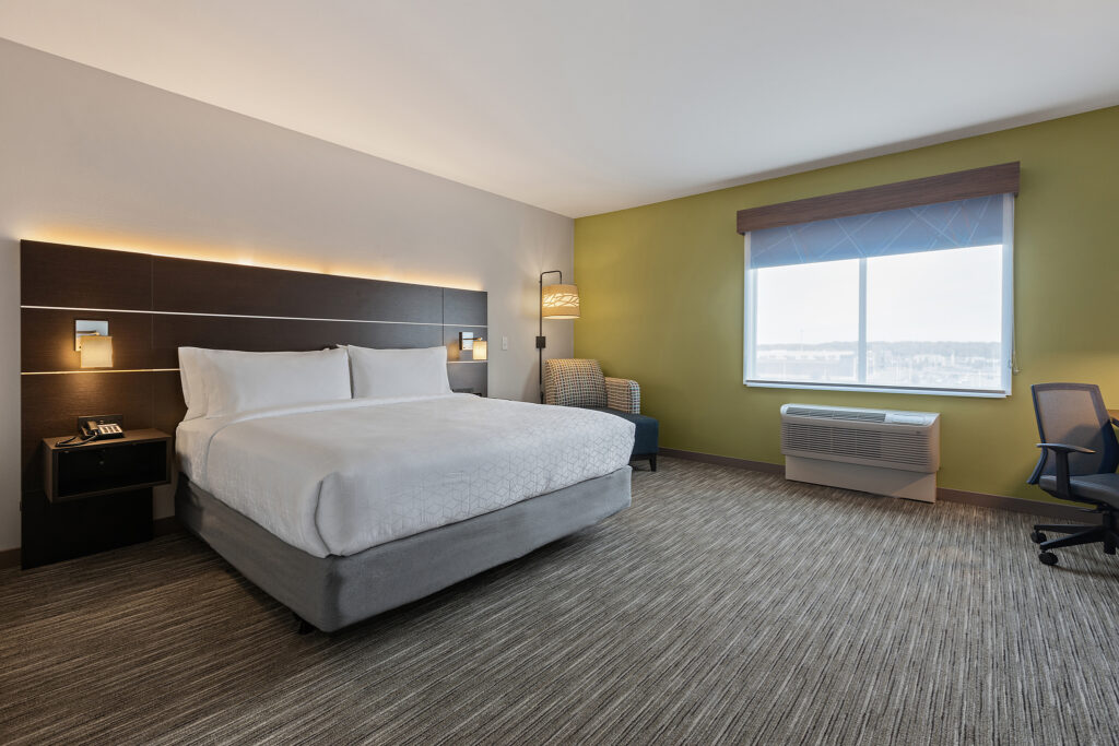 A modern Holiday Inn Express hotel room in Wisconsin features a large bed with crisp white bedding and a wooden headboard. A nightstand holds a phone, while an armchair with a lamp and a desk chair complete the space. Enjoy views through the window, framed by beige and green walls.