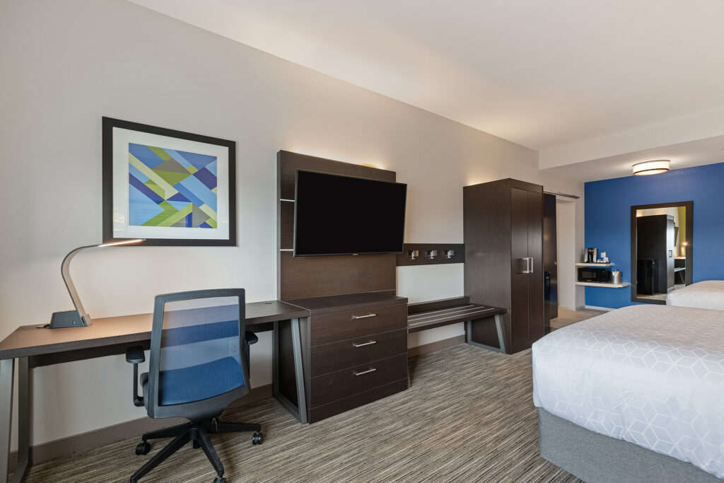 Experience a stay at a modern hotel room at Holiday Inn Express in Wisconsin, featuring two beds, a wall-mounted TV, and a desk equipped with a lamp and chair. Enjoy the artistic touch with abstract art and a blue accent wall, complemented by a large mirror and small beverage station for your convenience.