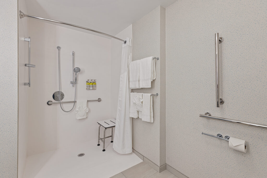 Located in Milwaukee, our accessible bathroom at Holiday Inn Express offers a walk-in shower with a bench, grab bars, and handheld showerhead for your convenience. Towels hang neatly on rods, complemented by a mounted toilet paper holder. The space is bright and impeccably clean.