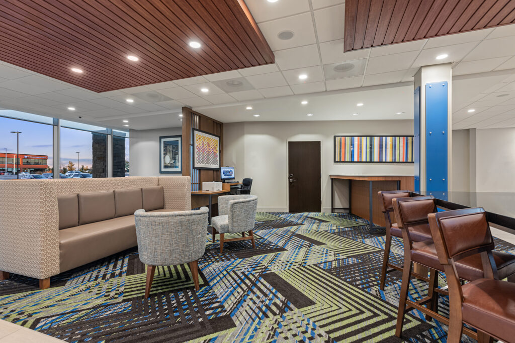 A modern lounge area with patterned carpet features a row of bar stools, two cushioned chairs, and a sofa. Decorated with colorful abstract art and large windows showcasing views of Wisconsin, the well-lit space is contemporary—typical of Holiday Inn Express's stylish design.