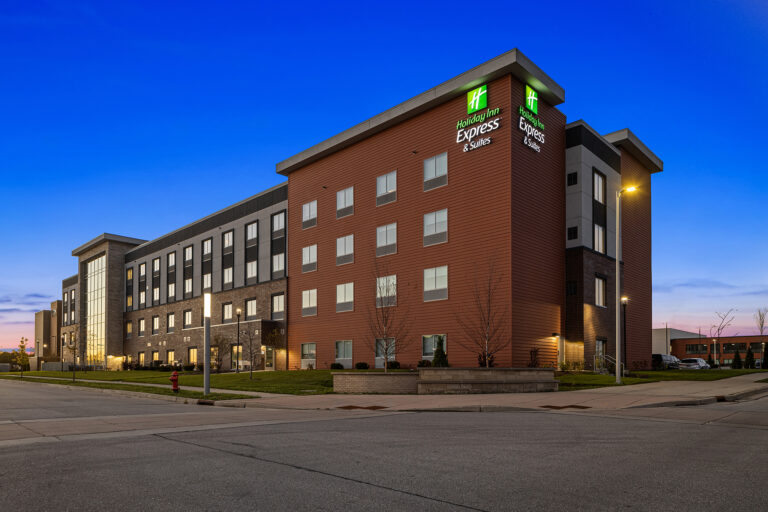 A modern hotel building, possibly an IHG property like Holiday Inn Express, features a blend of brick and dark paneling, standing prominently on a Milwaukee corner at dusk. The sky transitions from blue to orange, while streetlights illuminate the sidewalk and parking lot below.