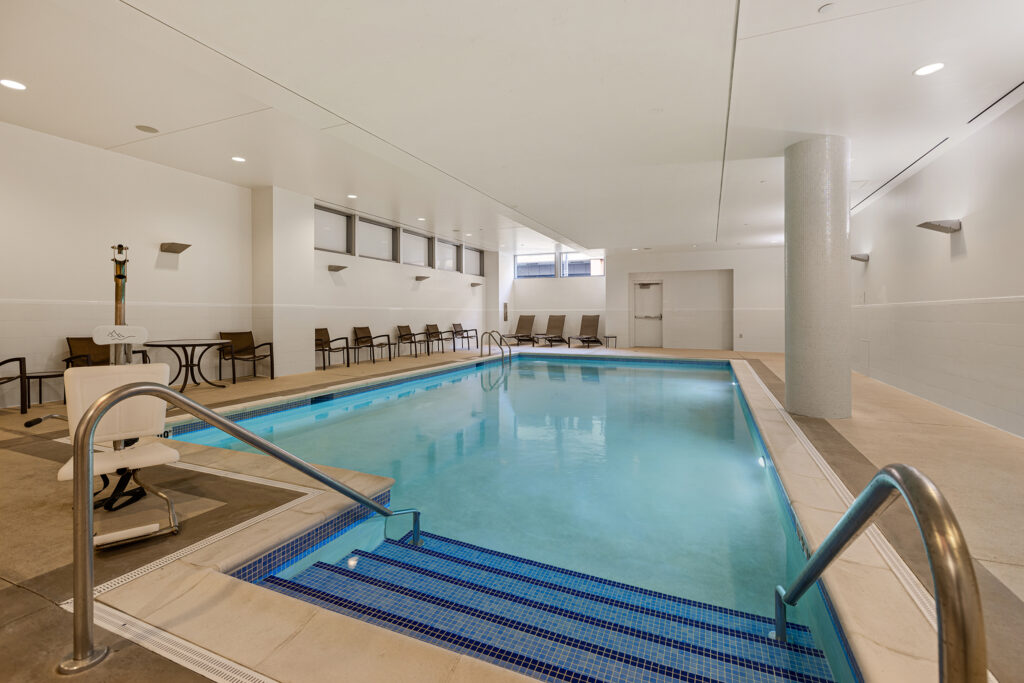 The indoor swimming pool at Hyatt Place Bloomington is nestled in a spacious room with white walls and ceiling. Ladders are at both ends, and lounge chairs line one side. A small table and decorative stand are visible on the left, while natural light filters through high windows.