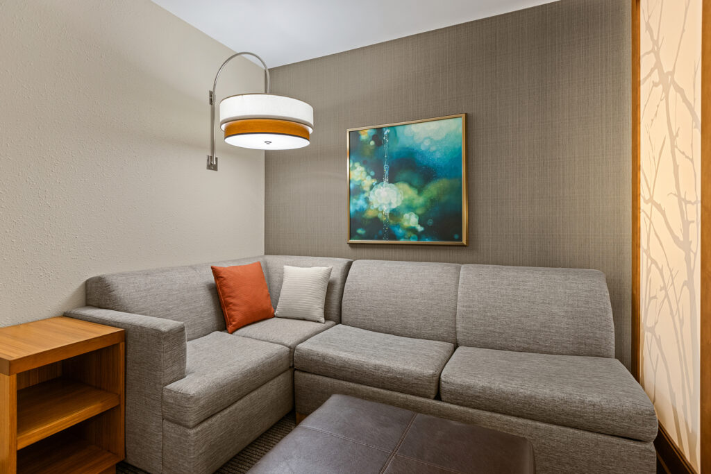 In a modern living room corner at the Bloomington Hyatt, a gray sectional sofa is adorned with one orange and one white pillow. Above the sofa, a contemporary art piece captivates. A circular ceiling lamp casts warm lighting, while a wooden side table stands to the left.