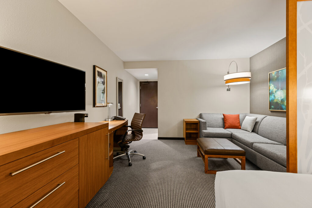 This modern hotel room in Bloomington, Illinois, offers a cozy atmosphere with neutral walls and carpeting. It features a large flat-screen TV on a wooden dresser, a workspace with a desk and chair, and a gray sectional sofa with a red cushion, perfect for relaxing under the stylish light fixture.
