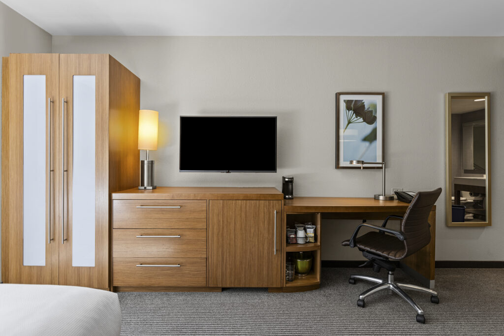 The modern hotel room at Hyatt Place showcases a wooden wardrobe and drawers on the left, with a flat-screen TV above. A desk with a chair and lamp complements the space. Wall art and a mirror enhance the light-colored walls, while Bloomington's charm is echoed in the carpeted floor.
