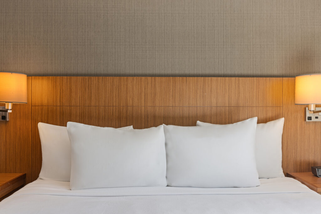 A neatly made bed with three large white pillows against a wooden headboard evokes an inviting atmosphere at the Hyatt in Bloomington, Illinois. Two wall-mounted lamps with beige shades cast warm light on the textured wallpaper behind the bed.