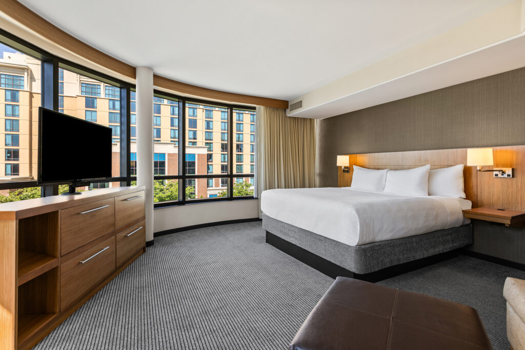Modern hotel room at Hyatt Place, featuring large windows, a king-sized bed, wall-mounted lamps, and a wooden dresser with a TV. The corner view showcases the vibrant streets of Bloomington and adjacent buildings, enhancing natural light effortlessly.