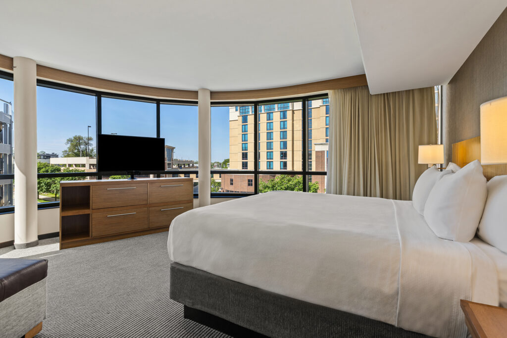 A spacious Hyatt Place hotel room in Illinois features a king-sized bed with white linens, a wooden dresser supporting a flat-screen TV, and large windows offering views of city buildings. Neutral-toned walls and carpet complement the decor, with a lamp mounted beside the bed.