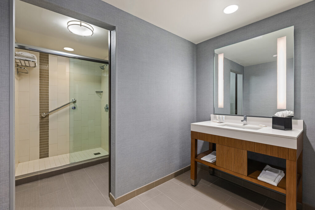 A modern bathroom at Hyatt Place Bloomington features a walk-in shower with a glass door, rainfall showerhead, and towel shelves. A wooden vanity with a large mirror and sink adorns the right side, all set against light gray walls and tile flooring.