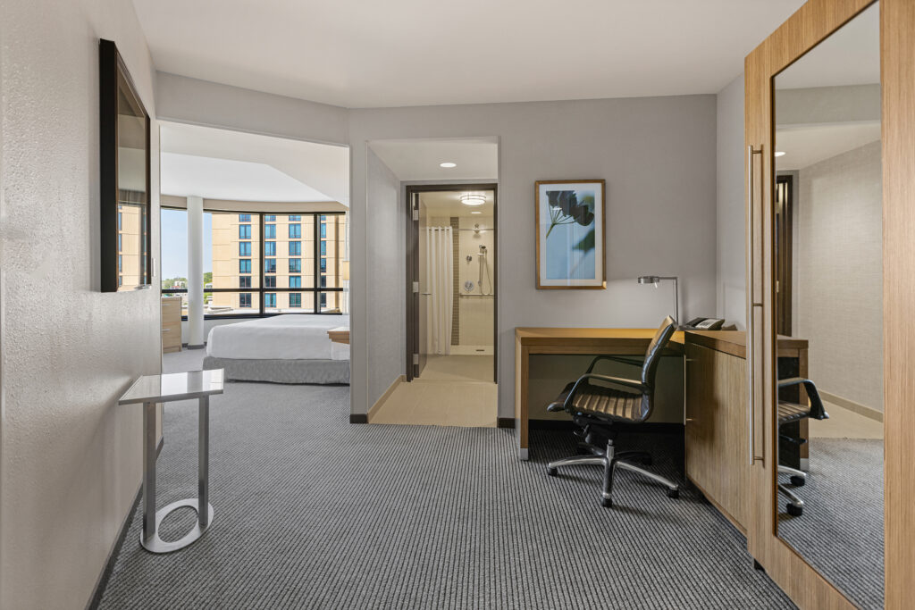 A modern room at Hyatt Place Bloomington offers a stunning cityscape view. It features a large bed, a work desk with chair, and a wall-mounted mirror. The bathroom boasts a glass shower, while the window bathes the stylish space in natural light.
