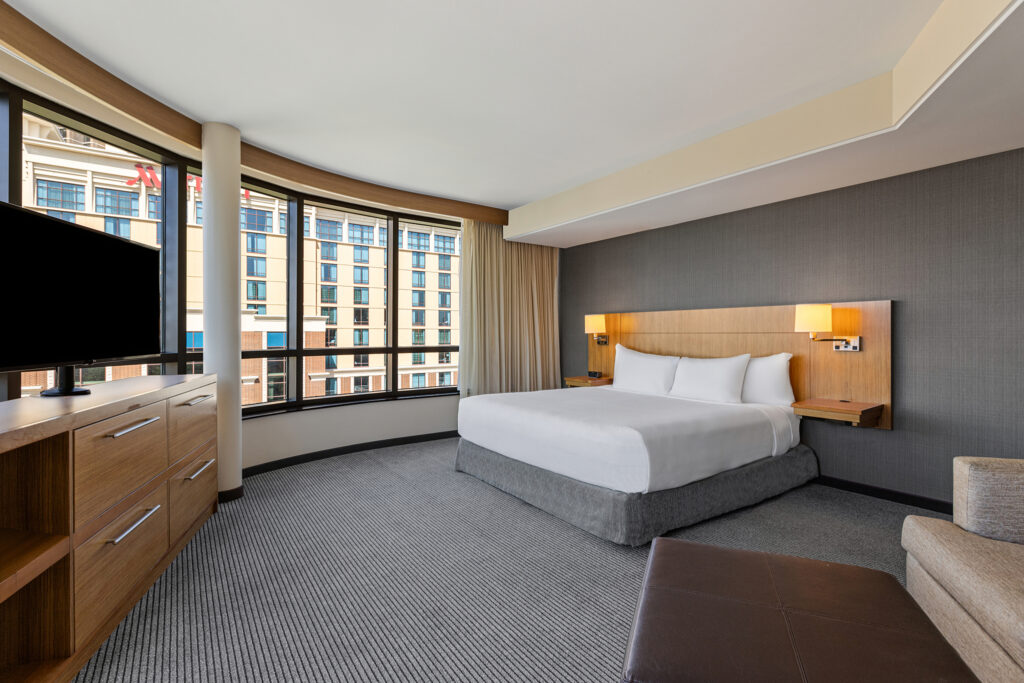 The modern hotel room at Hyatt Place Bloomington, Illinois, features a large bed, wall-mounted reading lights, and a flat-screen TV atop a wooden dresser. Large windows provide views of the cityscape. A gray armchair sits near the bed on a plush carpeted floor.