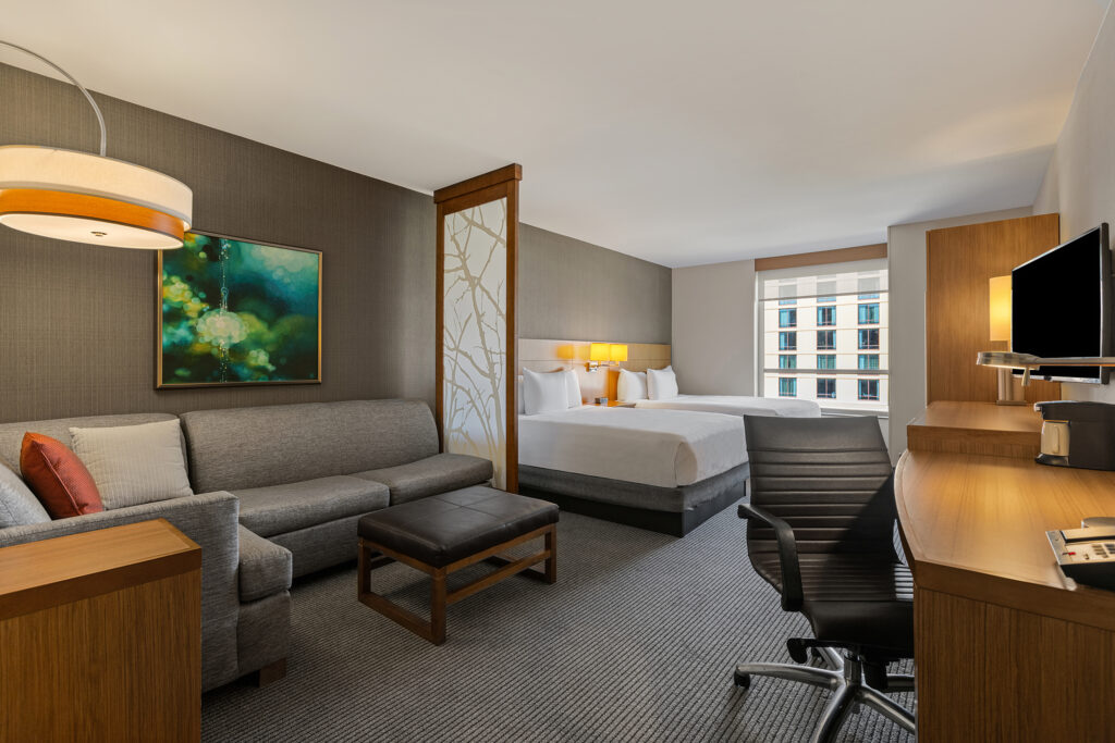 A modern hotel room at the Hyatt in Bloomington, Illinois, features a king-size bed, a gray sofa with a cushion, a coffee table, and a work desk with a chair. The wall-mounted TV, large window, ambient lighting, and art on the wall create an inviting atmosphere.