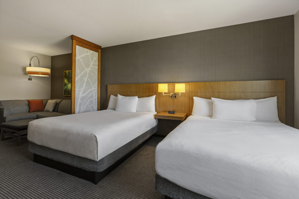 A modern Hyatt hotel room in Bloomington, Illinois, featuring two neatly made double beds with a nightstand and lamp between them. A sectional sofa graces the corner, while neutral-toned walls and carpeted floors create a cozy atmosphere. An abstract art piece adorns one wall.