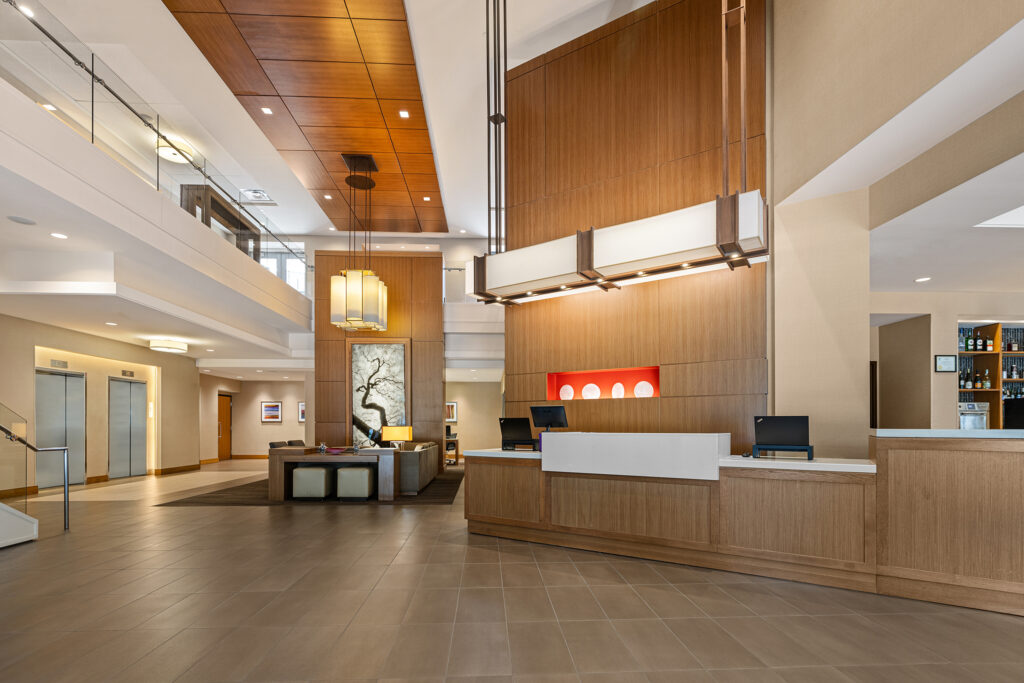 The spacious lobby of Hyatt Place Bloomington, Illinois, boasts modern decor with a large wooden reception desk, warm lighting, and a seating area. A framed artwork adorns the wall, while a staircase is visible to the left.