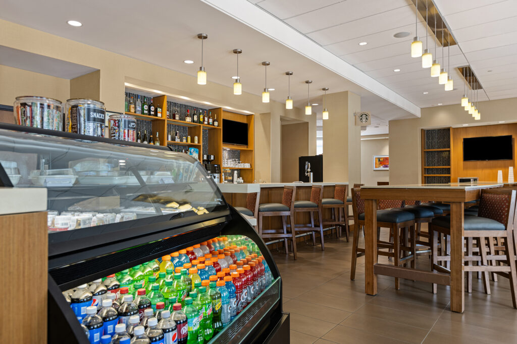 A modern cafe at the Hyatt in Bloomington features a refrigerated display of beverages and snacks. Behind, there's a bar area with cushioned stools, bottles on shelves, all set within a well-lit interior featuring ceiling lights and a flat-screen TV on the wall.