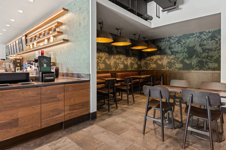 The interior of this modern coffee shop at Hyatt Place Bloomington boasts wooden counters, a hexagonal tile design, and a floral-patterned wall. The seating area features dark pendant lights with chairs and tables set against leafy wallpaper, creating a cozy atmosphere.