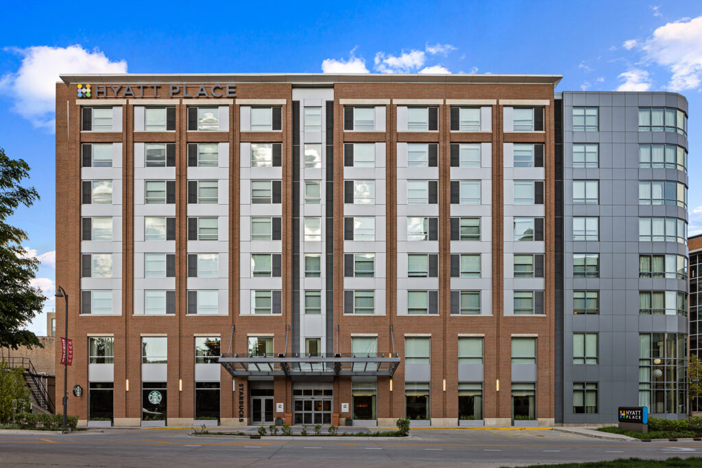 A multi-story Hyatt Place in Bloomington showcases a blend of brick and glass facade. The entrance boasts a modern canopy, complemented by the familiar Starbucks sign. Under the clear Illinois sky, with a few clouds, trees and a sidewalk enhance the inviting foreground.